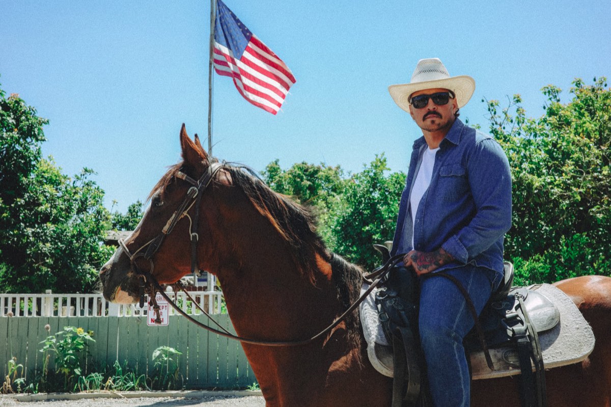 A man in a straw cowboy hat and denim shirt sits on a horse, wearing COE Bandit sunglasses, in front of a large American flag.